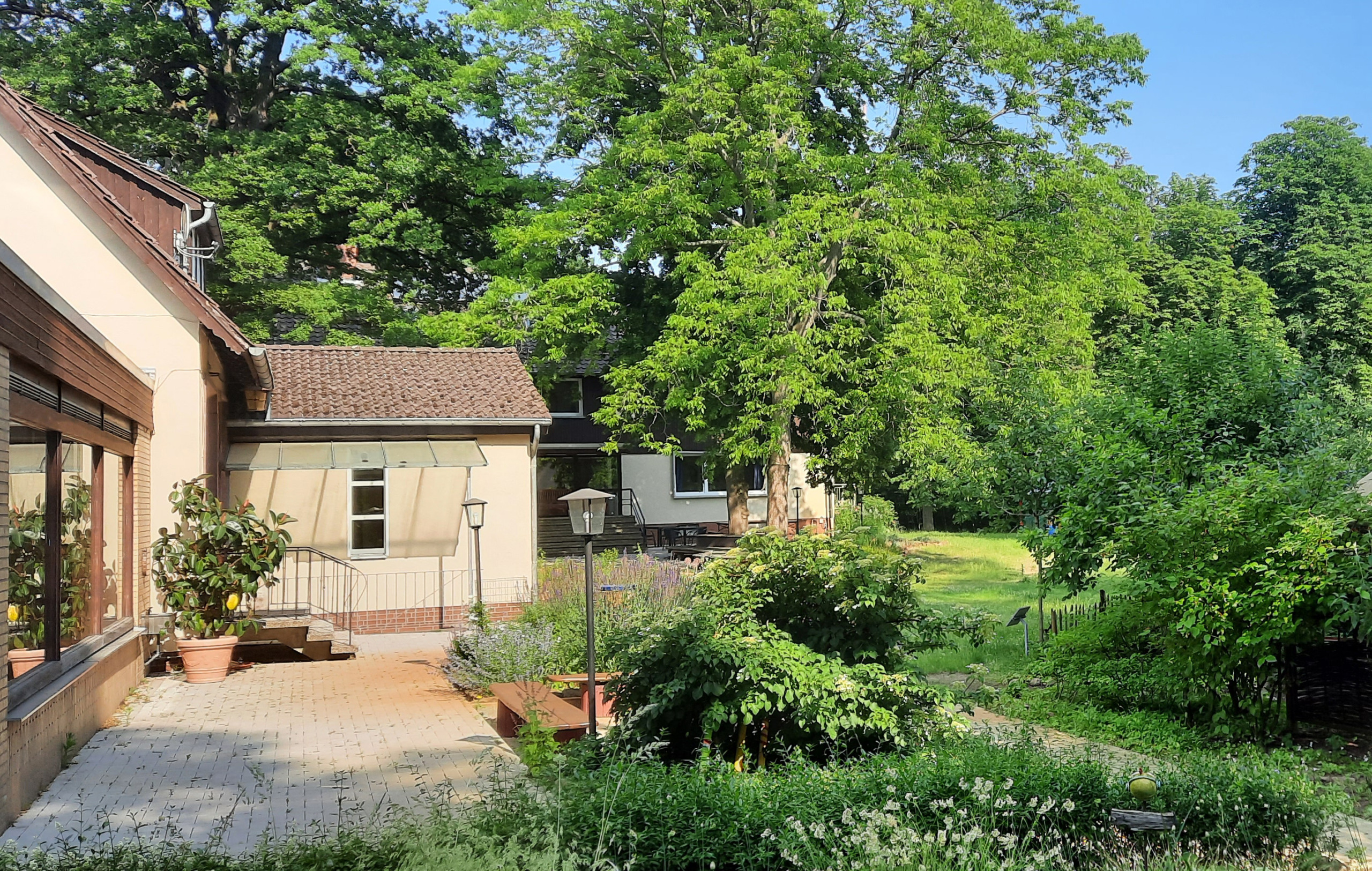 Zu sehen ist ein Teil des Naturfreundehauses mit Veranda, das umgeben ist von duftigen Wiesen, Büschen und Bäumen